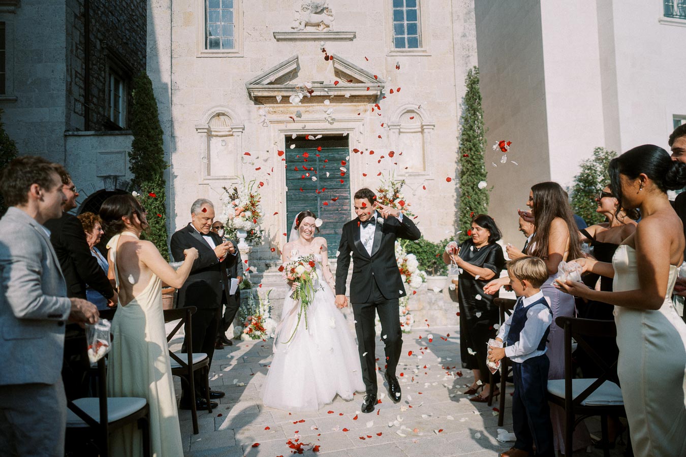 A joyful wedding ceremony outside a historic building, featuring a smiling bride in a white gown and groom in a black tuxedo, surrounded by guests showering them with rose petals.
