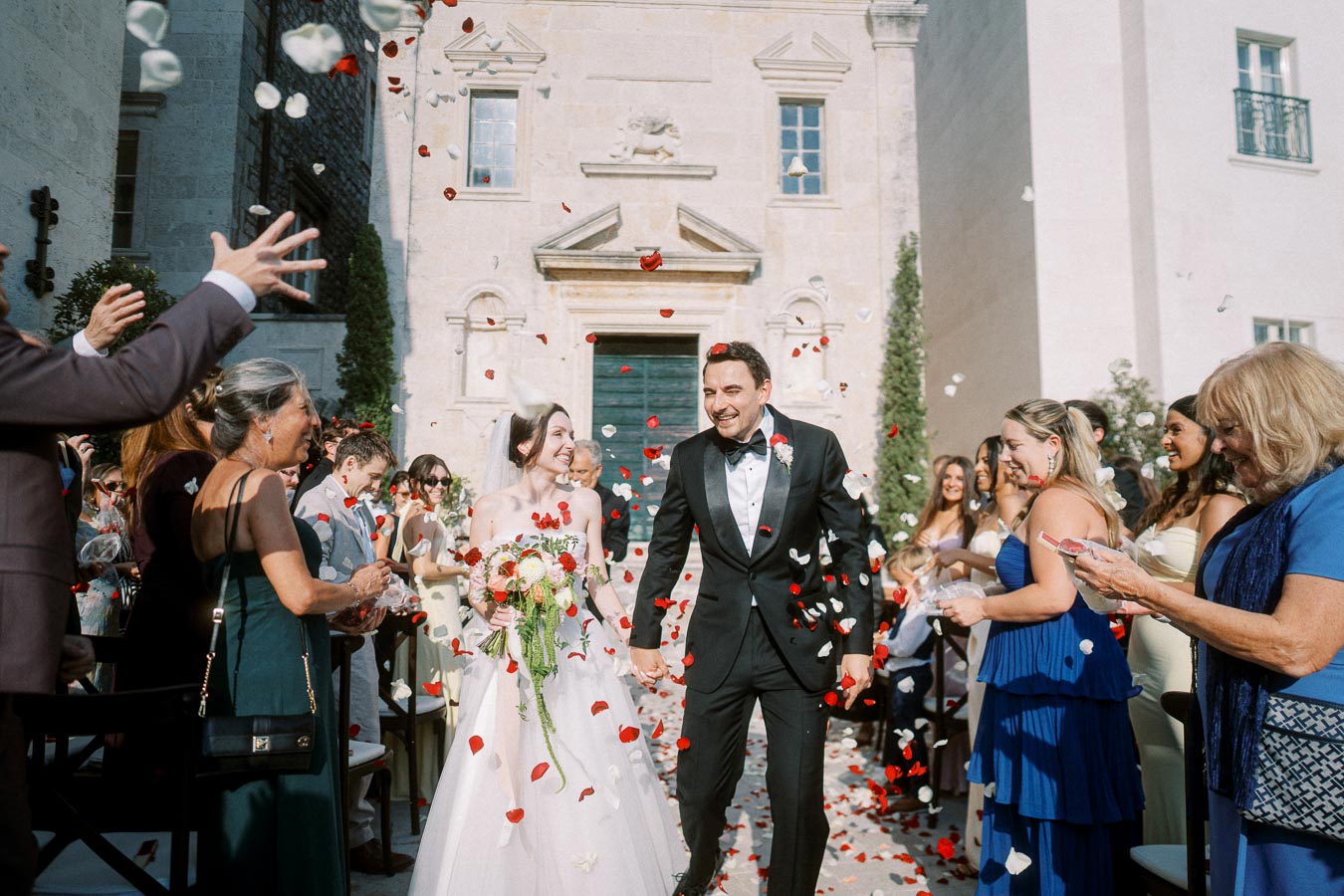 Bride and groom walking joyfully down the aisle, surrounded by guests throwing rose petals outside a historic church building.