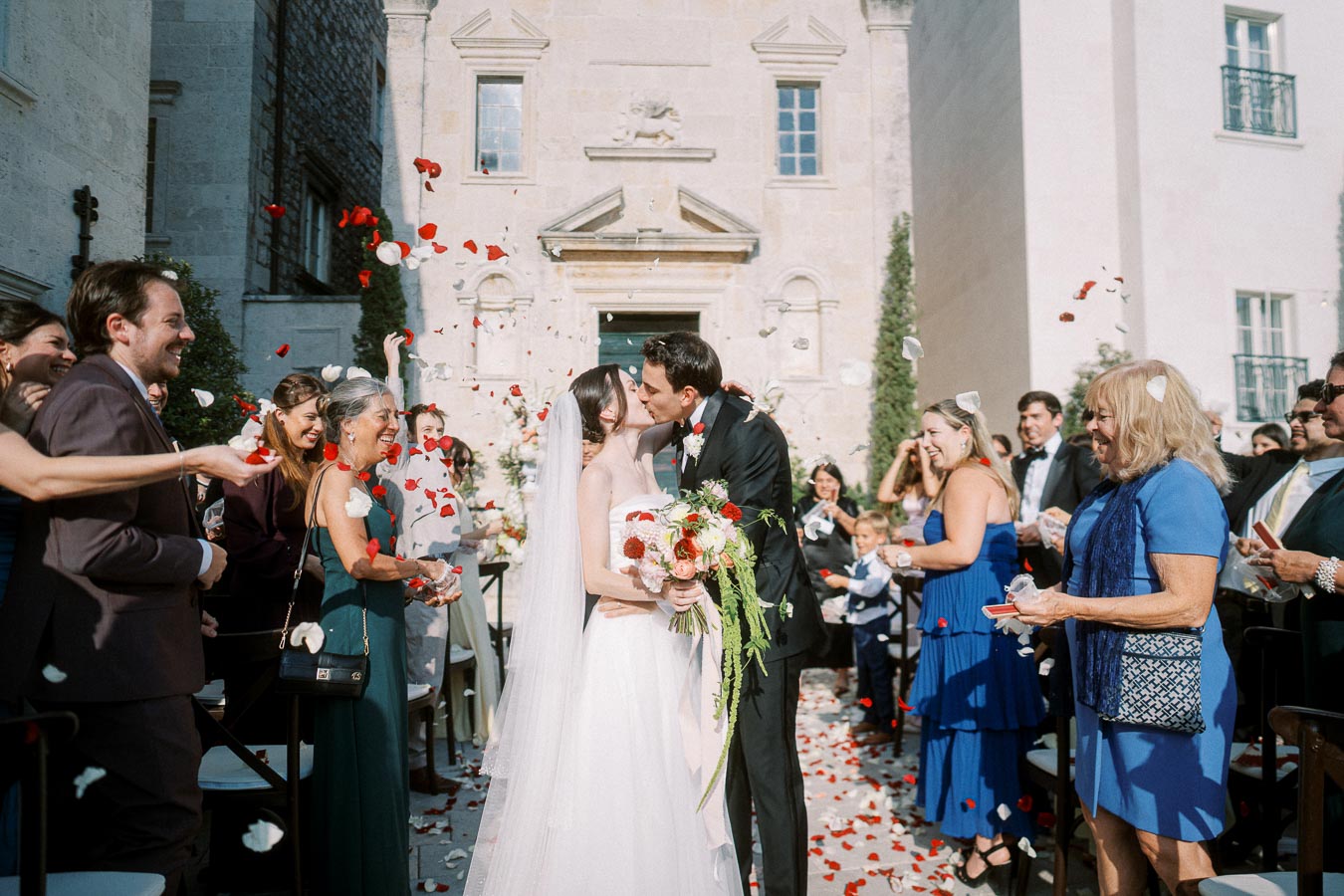 Bride and groom kissing at outdoor wedding ceremony surrounded by cheering guests throwing red and white rose petals, in front of a historic building.