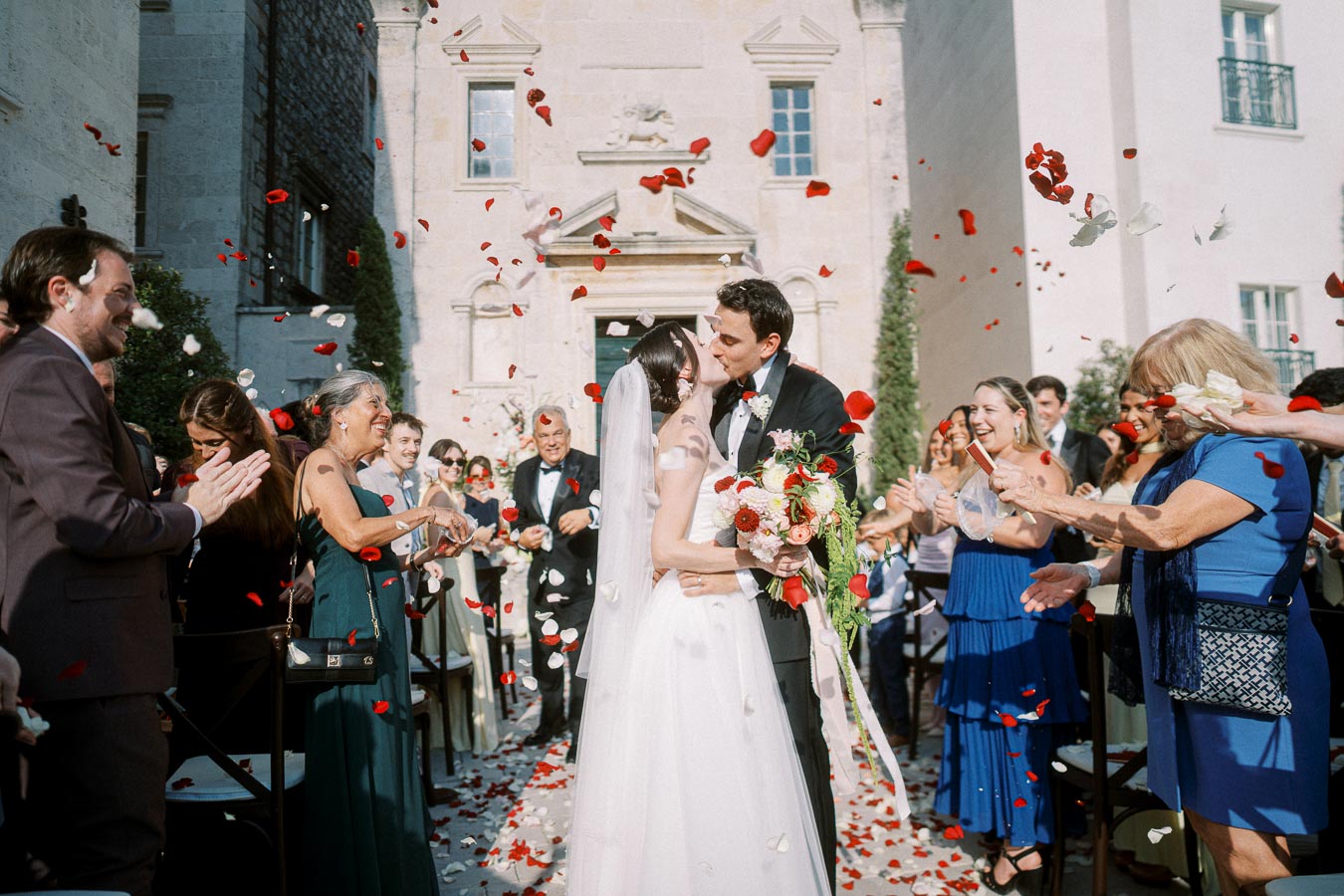 Bride and groom share a kiss surrounded by cheering guests and falling rose petals outside a historical stone building on their wedding day.