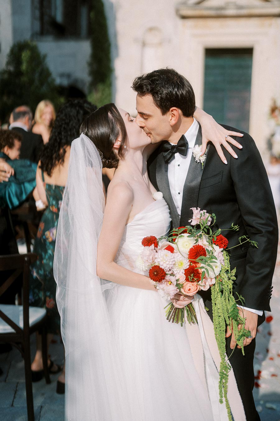 Elegant bride and groom sharing a kiss during outdoor wedding ceremony, bride holding vibrant floral bouquet, wearing white dress and veil, groom in black tuxedo.