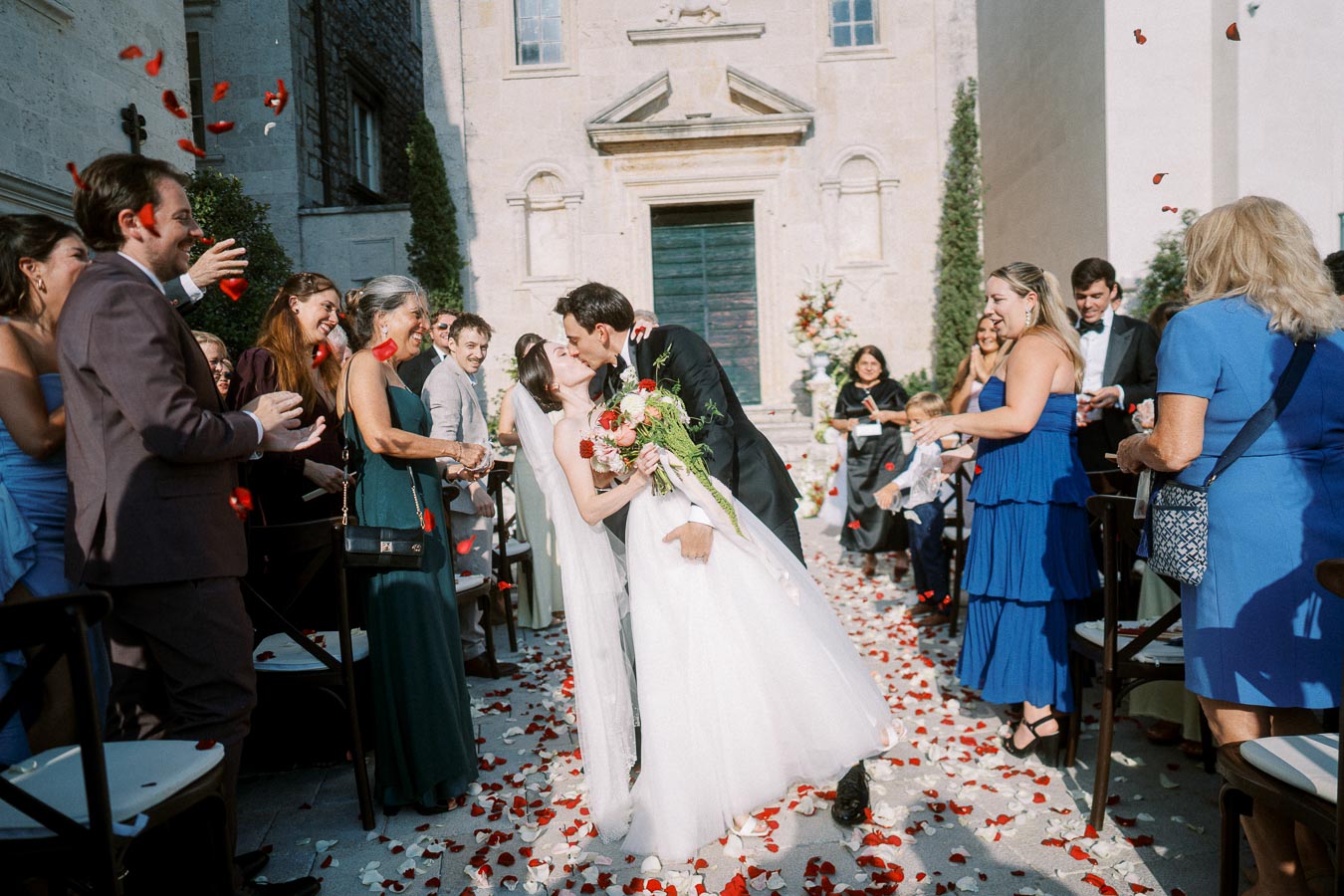 A bride and groom share a kiss surrounded by wedding guests in an outdoor ceremony, with rose petals scattered on the ground and in the air, creating a romantic atmosphere.