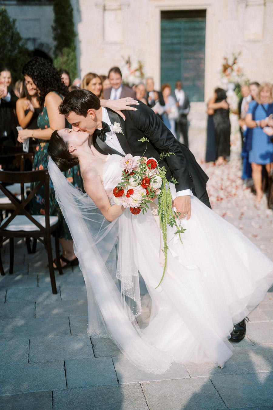 A newlywed couple shares a romantic kiss during their outdoor wedding ceremony, surrounded by guests. The bride wears a white gown with a long veil and holds a bouquet of red and white flowers, while the groom is in a black tuxedo.