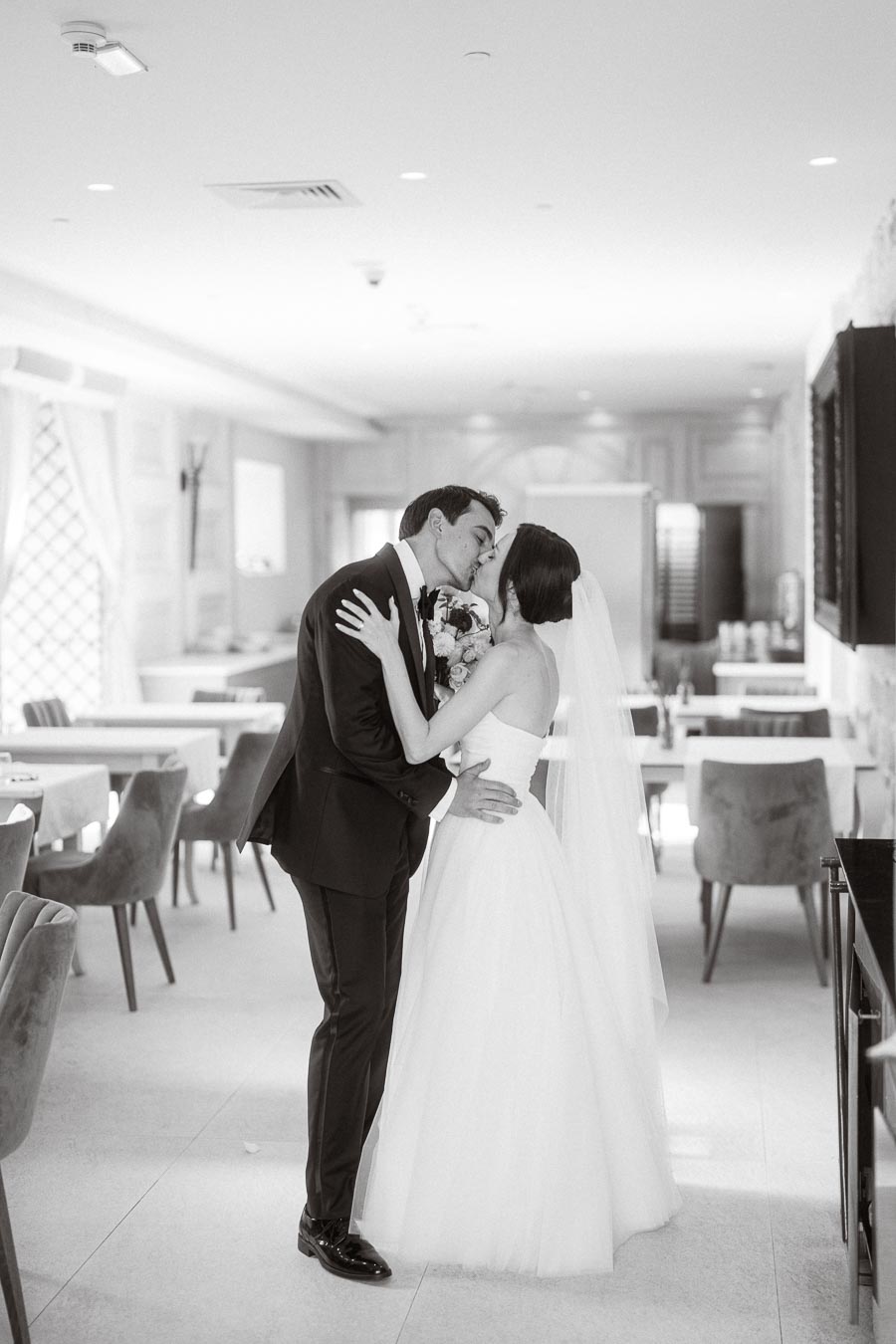 A bride and groom share a kiss in an elegant, modern venue, captured in black and white.