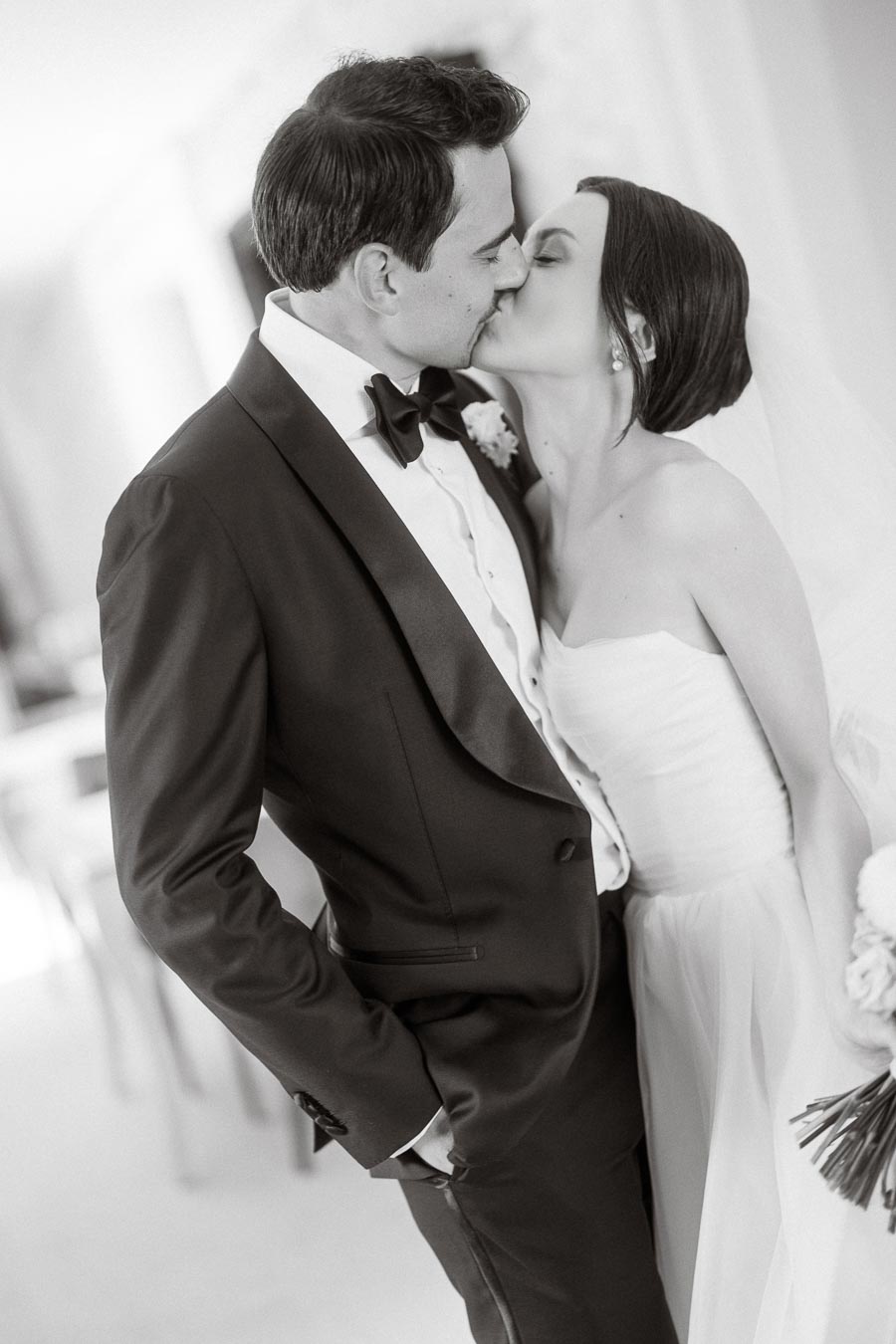 Black and white photo of a newlywed couple sharing a kiss, with the groom in a tuxedo and the bride in an elegant strapless gown, holding a bouquet.