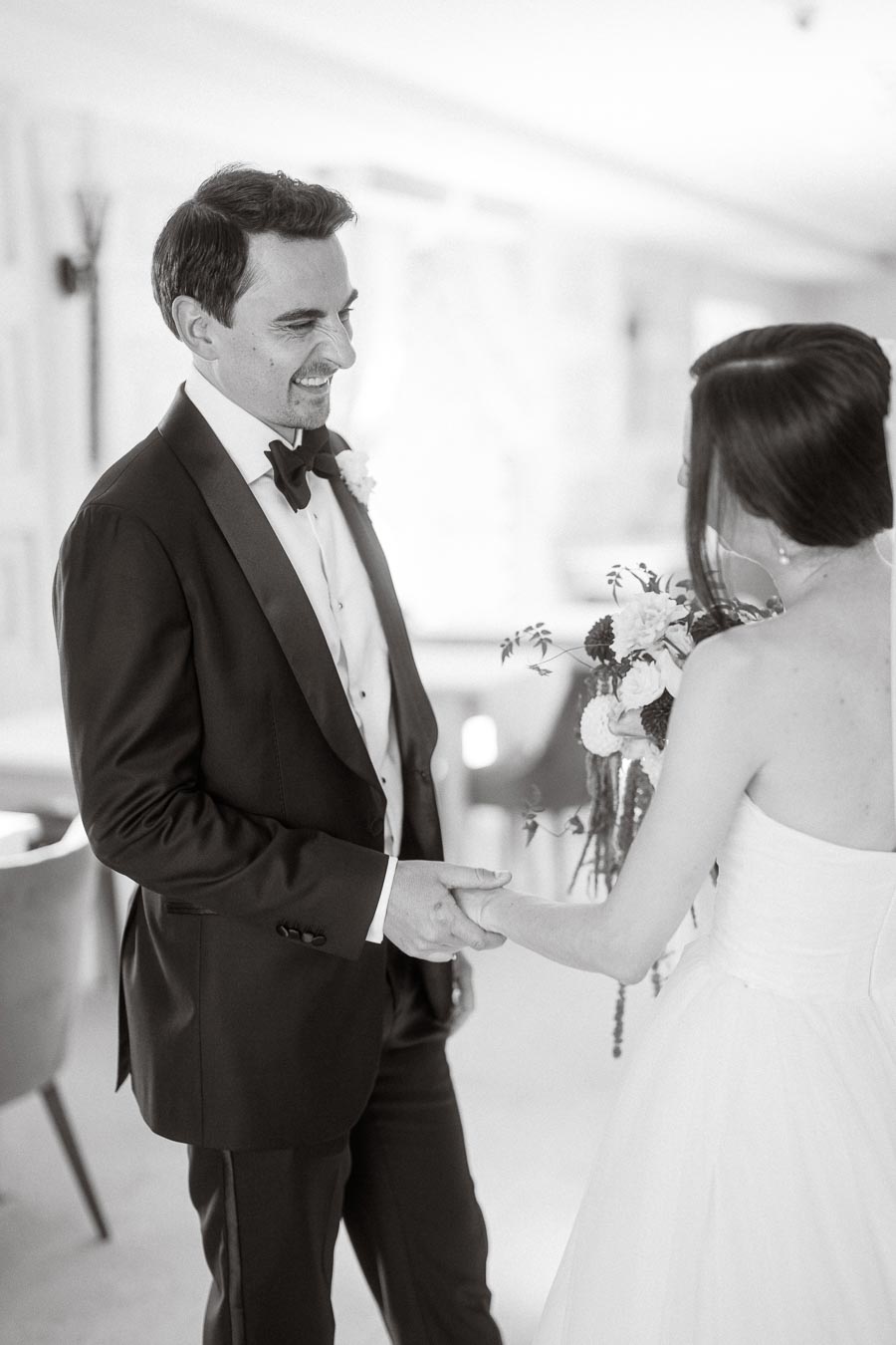 Black and white photo of a groom in a tuxedo smiling at his bride, who is wearing a strapless wedding dress and holding a bouquet, capturing a romantic moment on their wedding day.