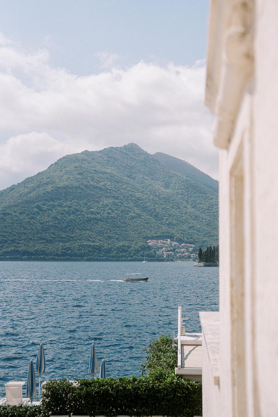 Scenic view of a tranquil bay with a boat gliding across the water, backed by lush green mountains under a partly cloudy sky, with beach umbrellas and a white building in the foreground.