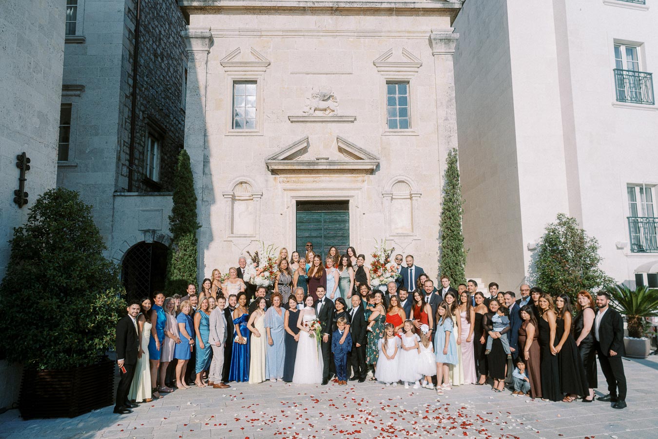 A large wedding group stands in front of an elegant stone building, featuring a mix of men, women, and children dressed in formal attire. The outdoor scene is decorated with scattered rose petals on the ground, and lush greenery flanks the building's entrance.
