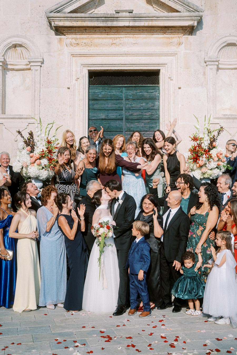 Wedding celebration with a large group of smiling guests outside a historic building, as a newlywed couple shares a kiss at the center, surrounded by loved ones in colorful attire and floral arrangements.