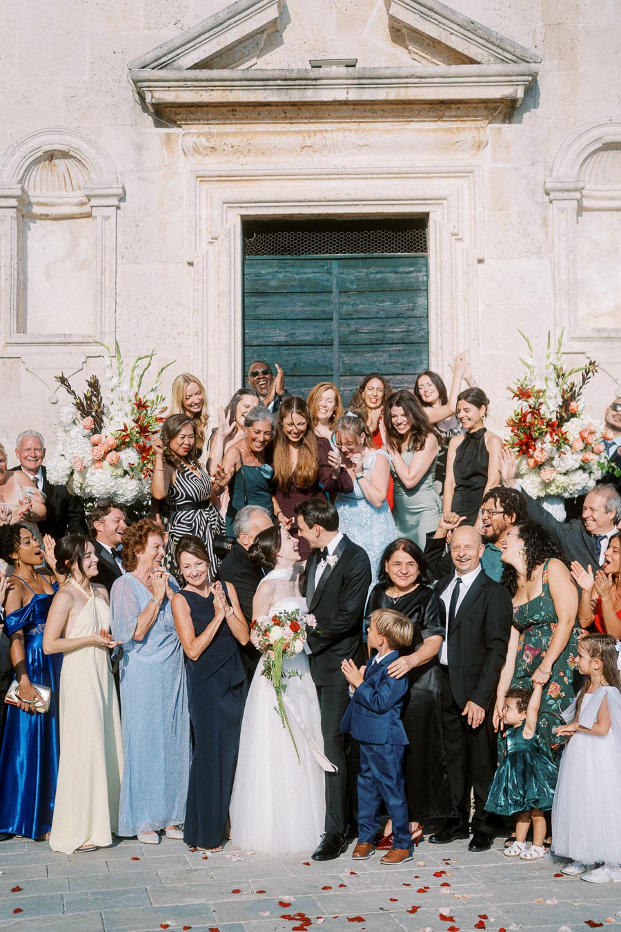 A large group of people celebrating a wedding outside a stone building, with a bride in a white dress and groom in a black suit surrounded by smiling guests, including men, women, and children, dressed in formal attire with floral decorations in the background.