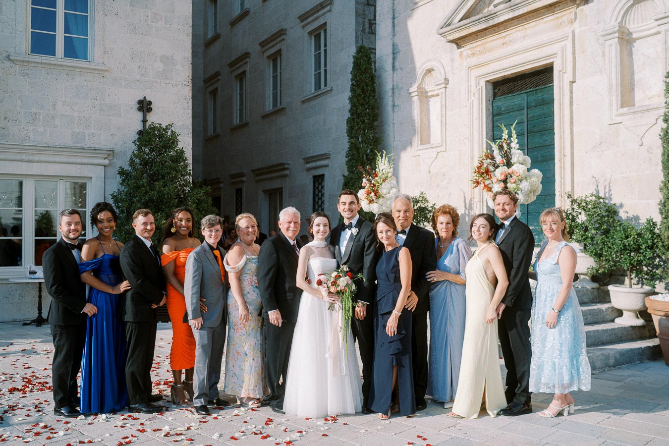 Wedding party posing outside a historic stone building, with bride, groom, and diverse group of guests smiling in formal attire, surrounded by floral arrangements.