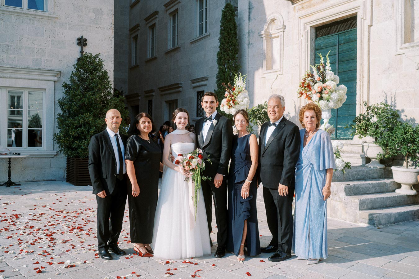 A wedding party posing for a photo outside a historic stone building, with a bride in a white gown and a groom in a black tuxedo surrounded by elegantly dressed family members, amidst scattered rose petals.