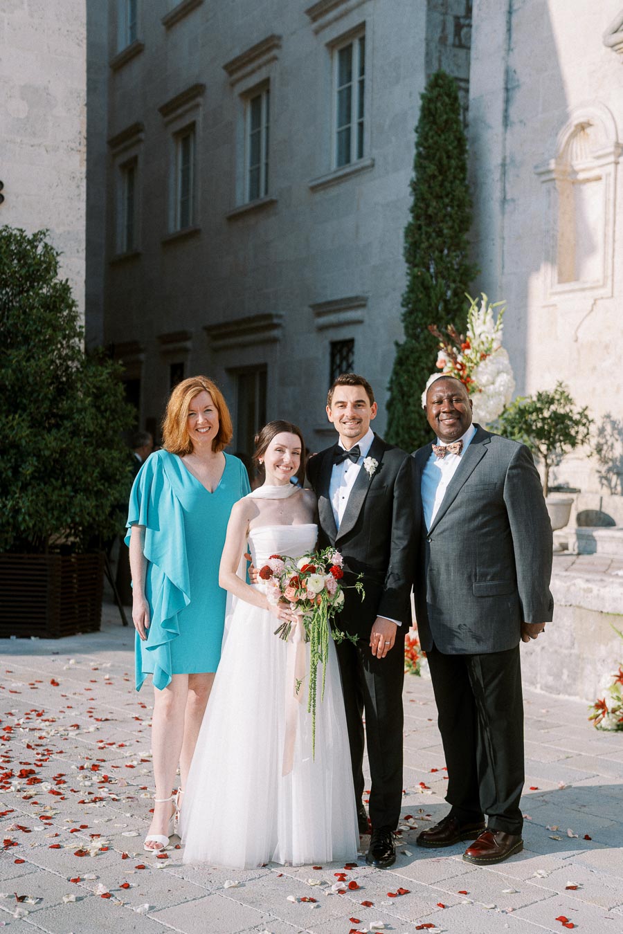 A bride in a white gown and groom in a tuxedo smile with two guests at an outdoor wedding. The woman in blue and man in a dark suit stand beside the couple, surrounded by scattered rose petals and a historic building backdrop.