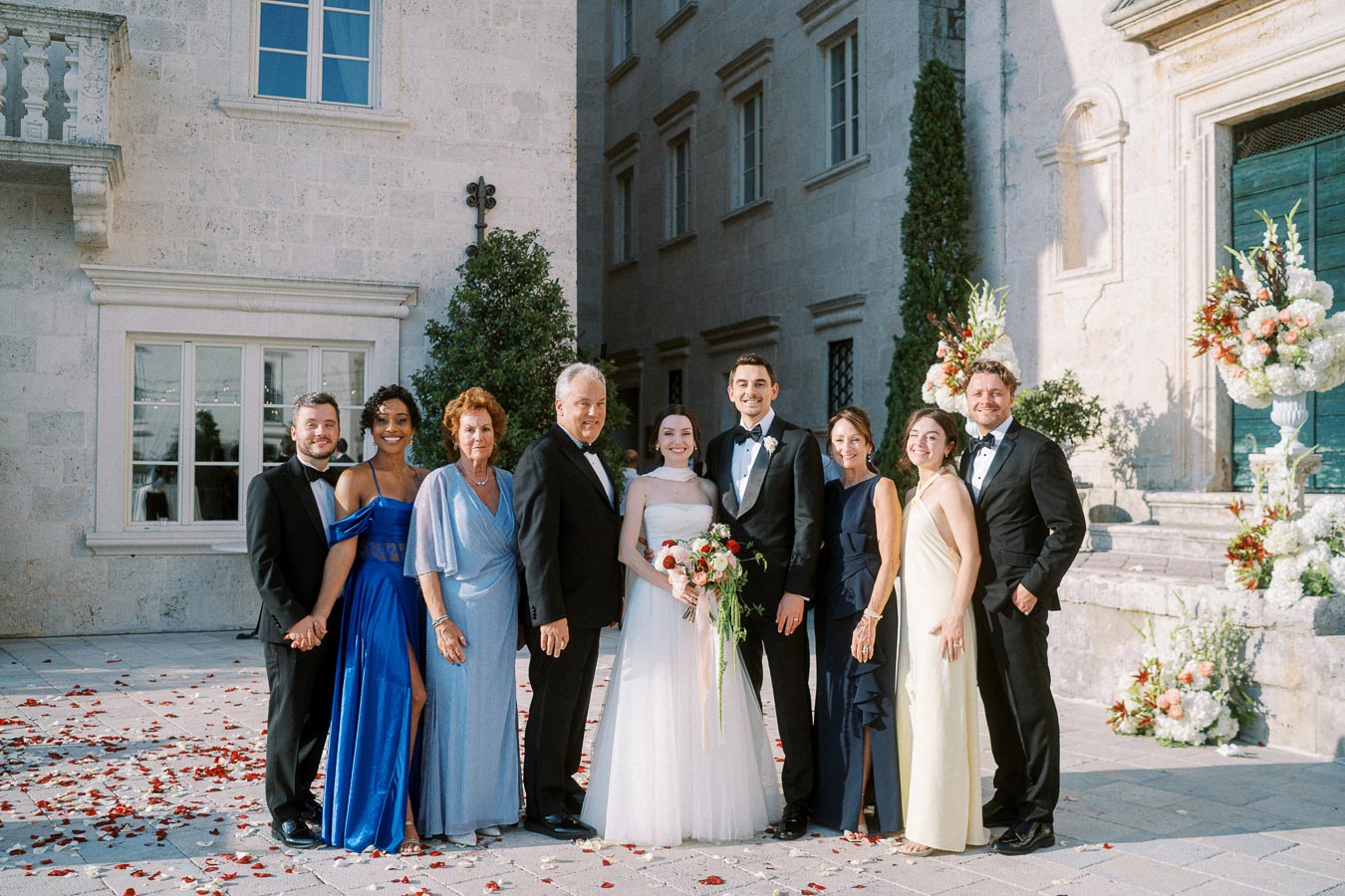 Wedding party posing for a group photo outside a historical building, featuring a bride in a white gown, groom in a tuxedo, and family members dressed in elegant formal attire, surrounded by floral decorations and rose petals.