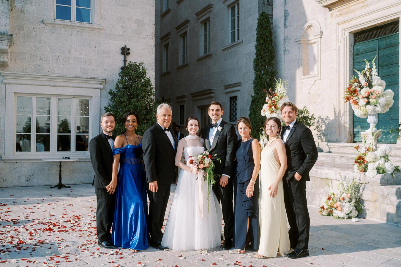 A wedding party poses together outside a beautiful stone building adorned with floral arrangements, featuring the bride in a white gown and groom in a tuxedo, surrounded by family and friends in formal attire on a sunlit day.