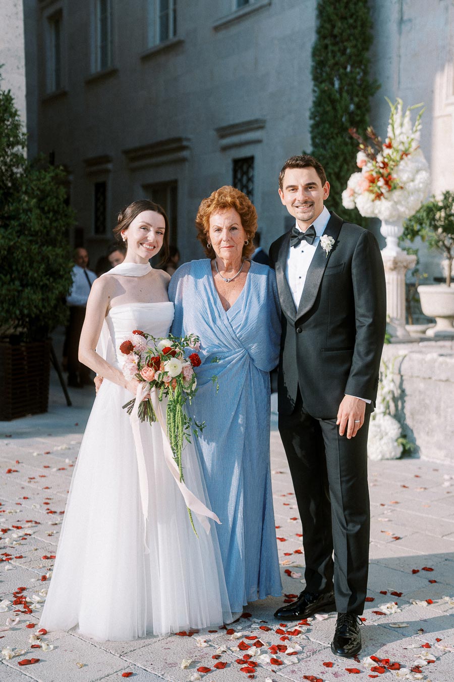 A bride in a white dress and bouquet stands with an elderly woman in a blue gown and a groom in a black tuxedo. They are posing outdoors on a sunny day with flower petals scattered on the ground and elegant stone architecture in the background.
