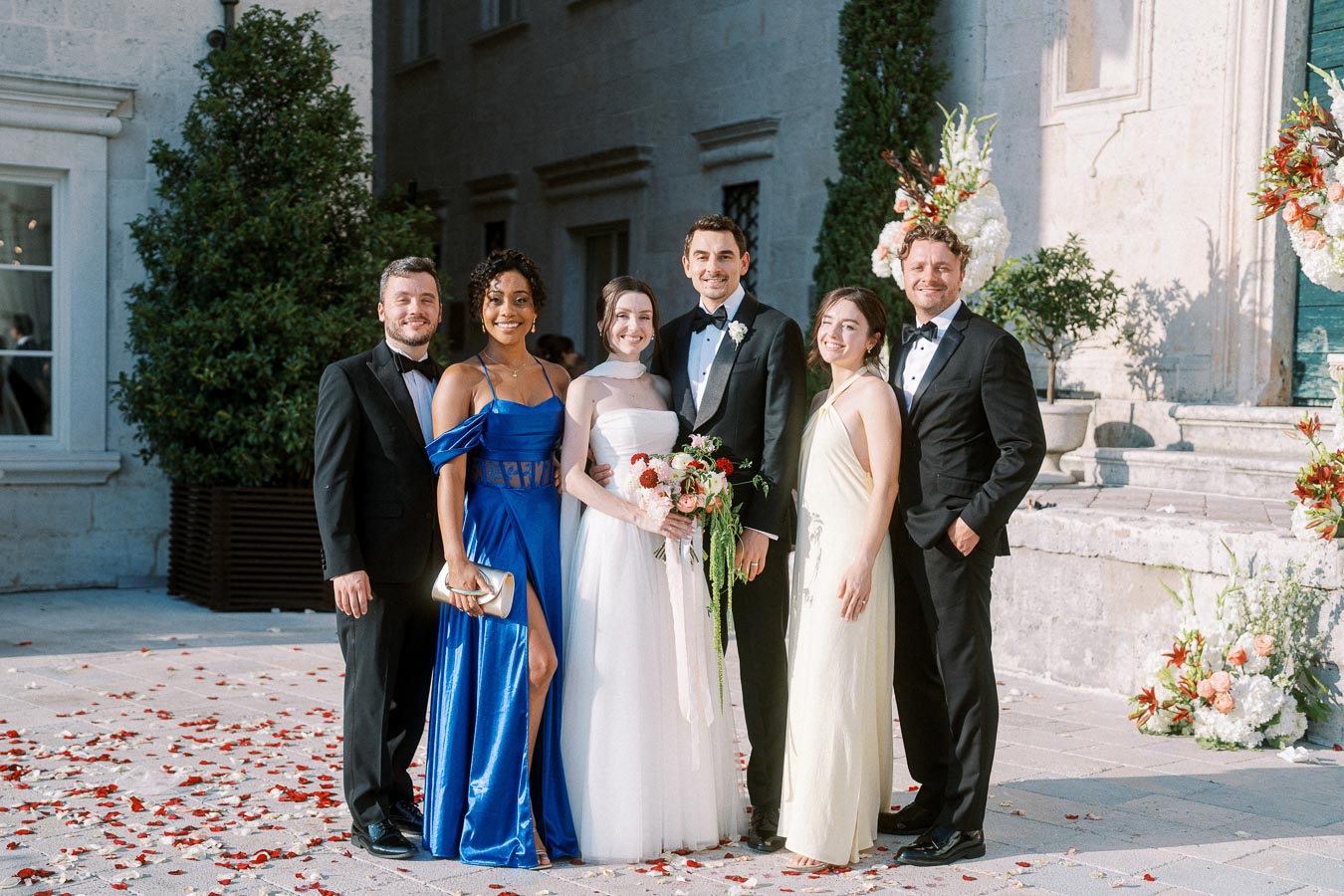 A wedding group portrait with six elegantly dressed people standing outdoors, featuring a bride in a white dress holding a bouquet and a groom in a tuxedo, surrounded by friends in formal attire, with a stone building and floral decorations in the background.