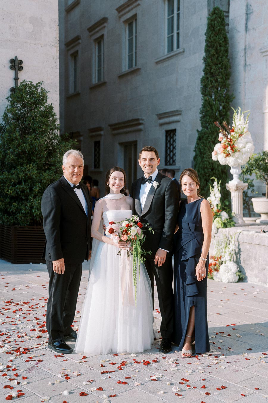 Wedding ceremony photo featuring a bride and groom smiling with two elegantly dressed relatives, outdoor venue adorned with flower petals, formal attire, celebration atmosphere.