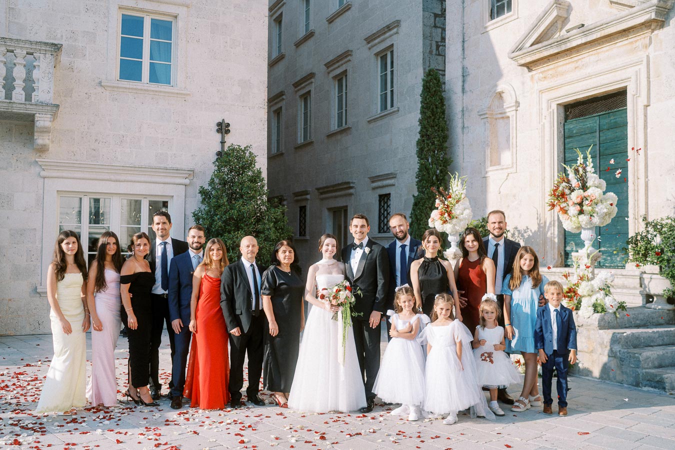 Wedding party group photo outside a historic building, featuring the bride and groom surrounded by elegantly dressed family and friends.