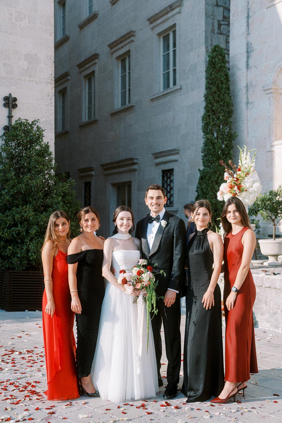 A wedding couple posing with four bridesmaids outside an elegant stone building, with scattered rose petals on the ground. The bride holds a vibrant bouquet, while the groom wears a classic black tuxedo. The bridesmaids are dressed in stylish red and black dresses.