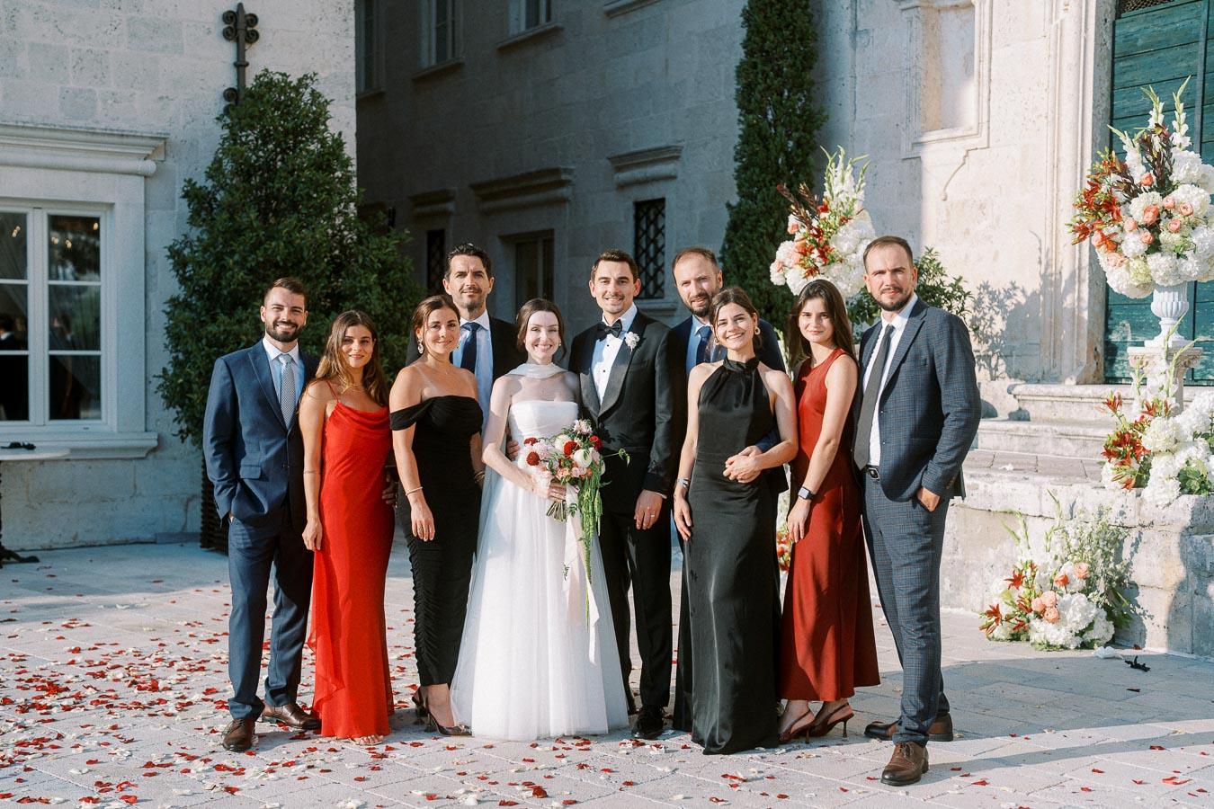 A group of elegantly dressed people posing for a wedding photo in a courtyard, with a bride in a white gown holding a bouquet and a groom in a tuxedo, surrounded by smiling guests in formal attire.