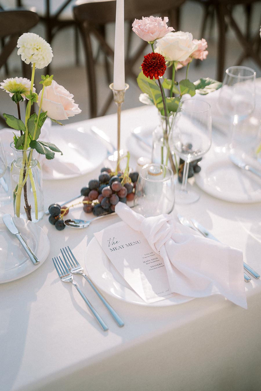Elegant wedding table setting with a white tablecloth, featuring a menu labeled The Meat Menu, a neatly folded napkin, and polished silverware. The arrangement is accented by colorful flowers in glass vases, red and white blooms, and a cluster of grapes, all positioned alongside wine glasses and a tall candle.