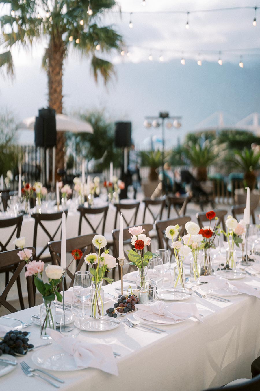 Outdoor wedding reception with elegant table settings featuring white tablecloths, glass plates, cutlery, and floral arrangements with colorful flowers. Grapes are placed on tables, and the setting is enhanced by a tropical palm tree and string lights overhead, creating a romantic atmosphere.