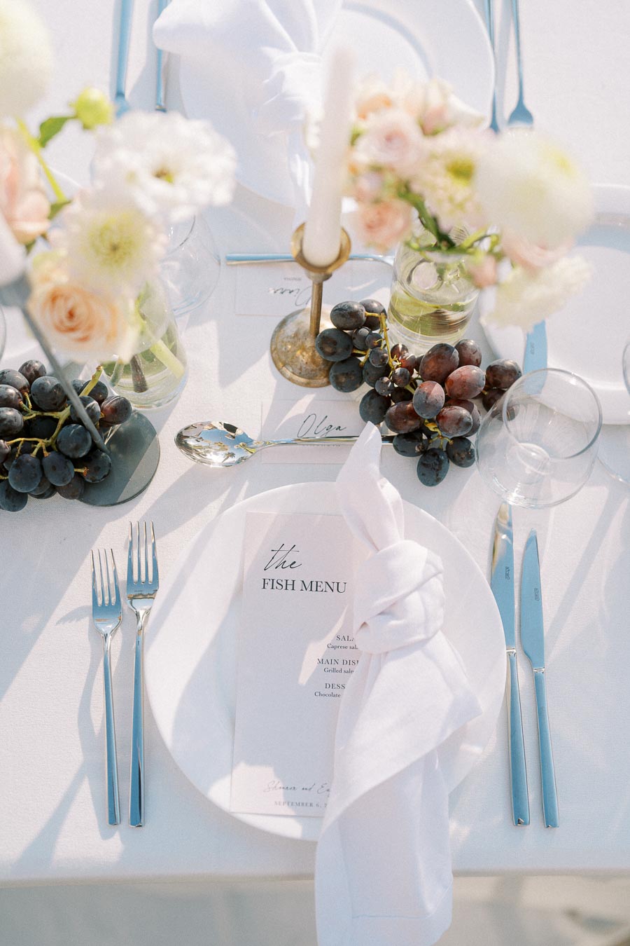 Elegant wedding table setting with white floral arrangements, grapes, and a fish menu on a white tablecloth; features silver cutlery and a delicate napkin fold.
