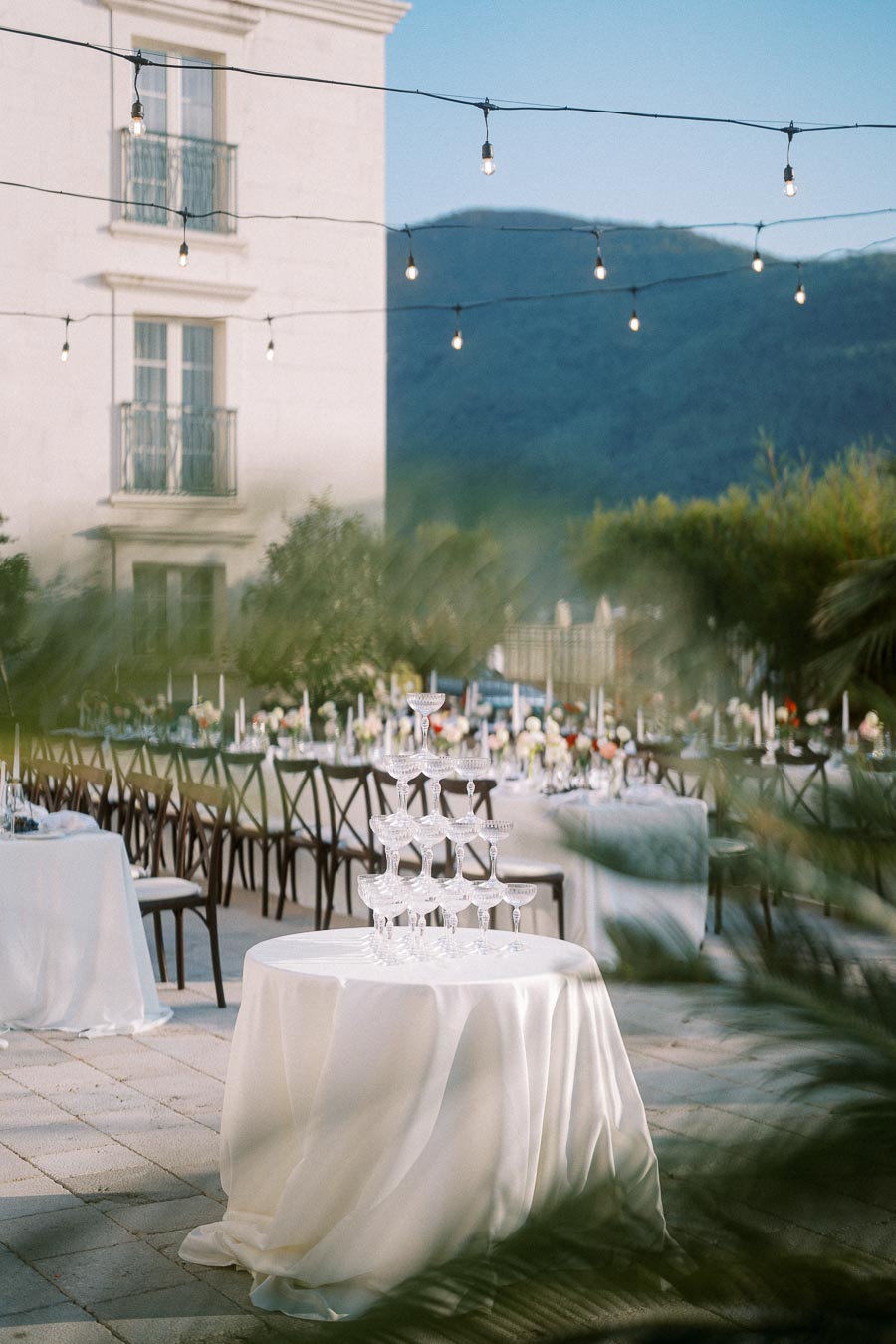 Outdoor wedding reception setting with elegantly decorated tables, string lights, and a champagne tower in focus, surrounded by scenic mountain views and a building in the background.