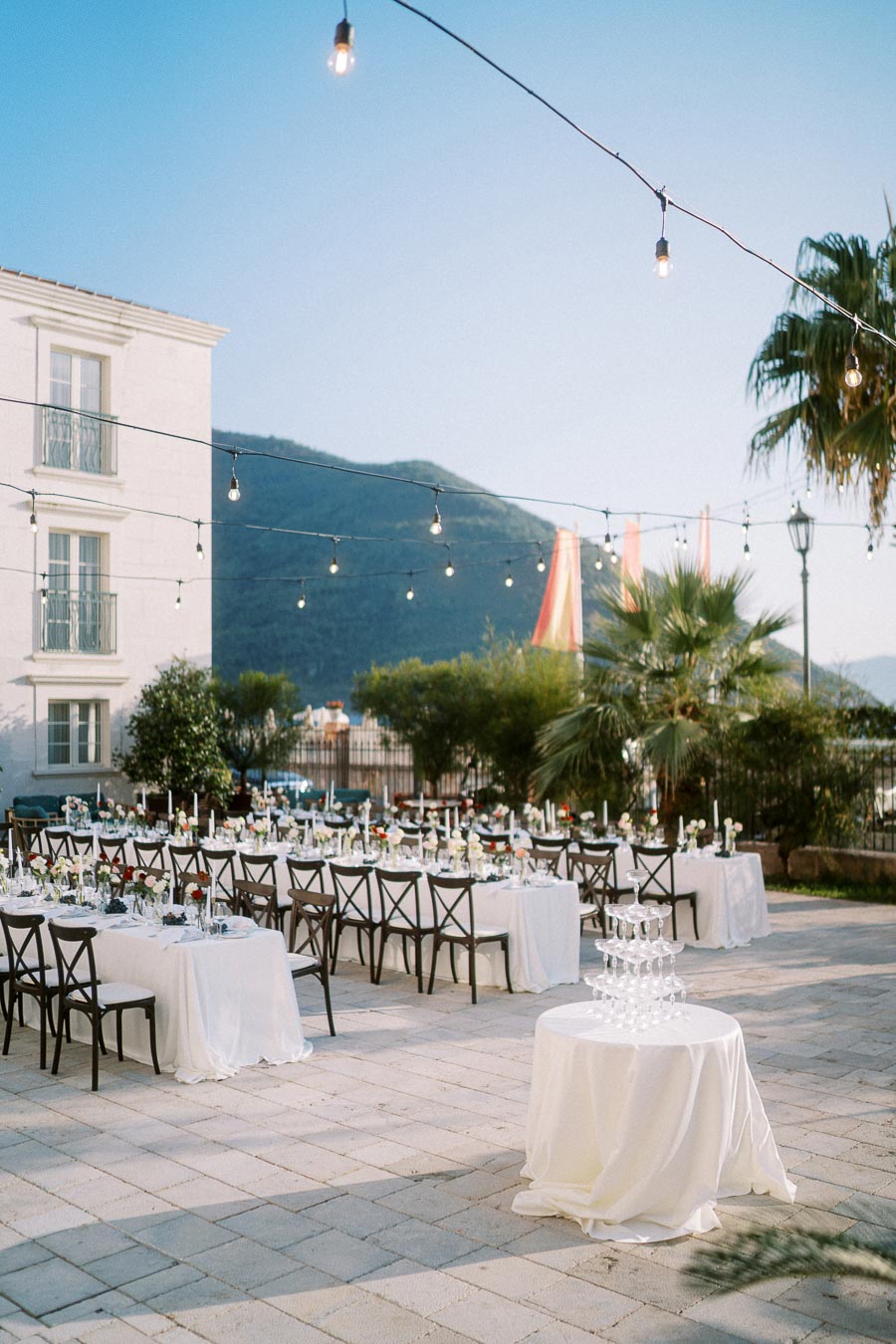 Outdoor wedding reception setup with elegantly decorated tables and chairs, adorned with white tablecloths and floral centerpieces. A champagne tower stands on a small round table, surrounded by string lights, trees, and a scenic mountain backdrop.