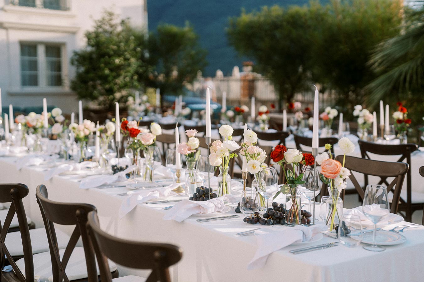 Elegant outdoor wedding reception with long tables adorned with floral centerpieces, white candles, and place settings, set against a lush garden backdrop.