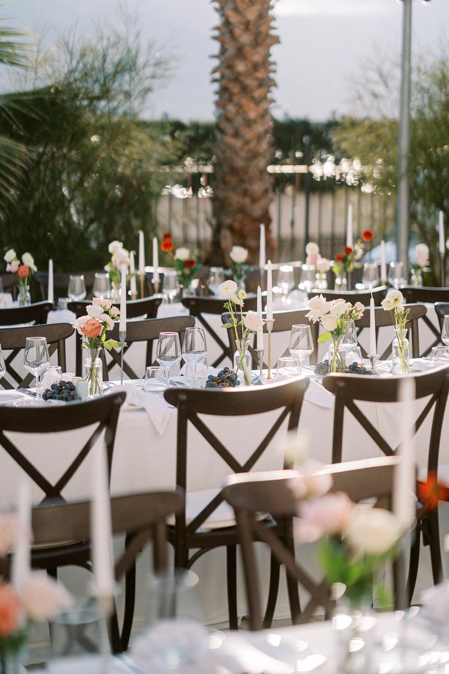 Elegant outdoor wedding reception setup with tables adorned with white tablecloths, floral centerpieces, and candles, surrounded by natural greenery and palm trees.