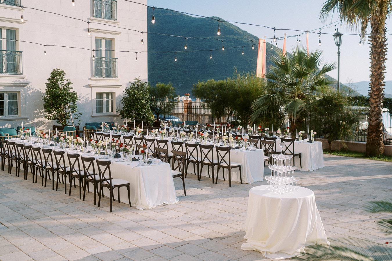 Outdoor wedding reception setup with elegantly decorated long tables under string lights, featuring floral centerpieces and a champagne tower, located in a scenic venue with mountains in the background.