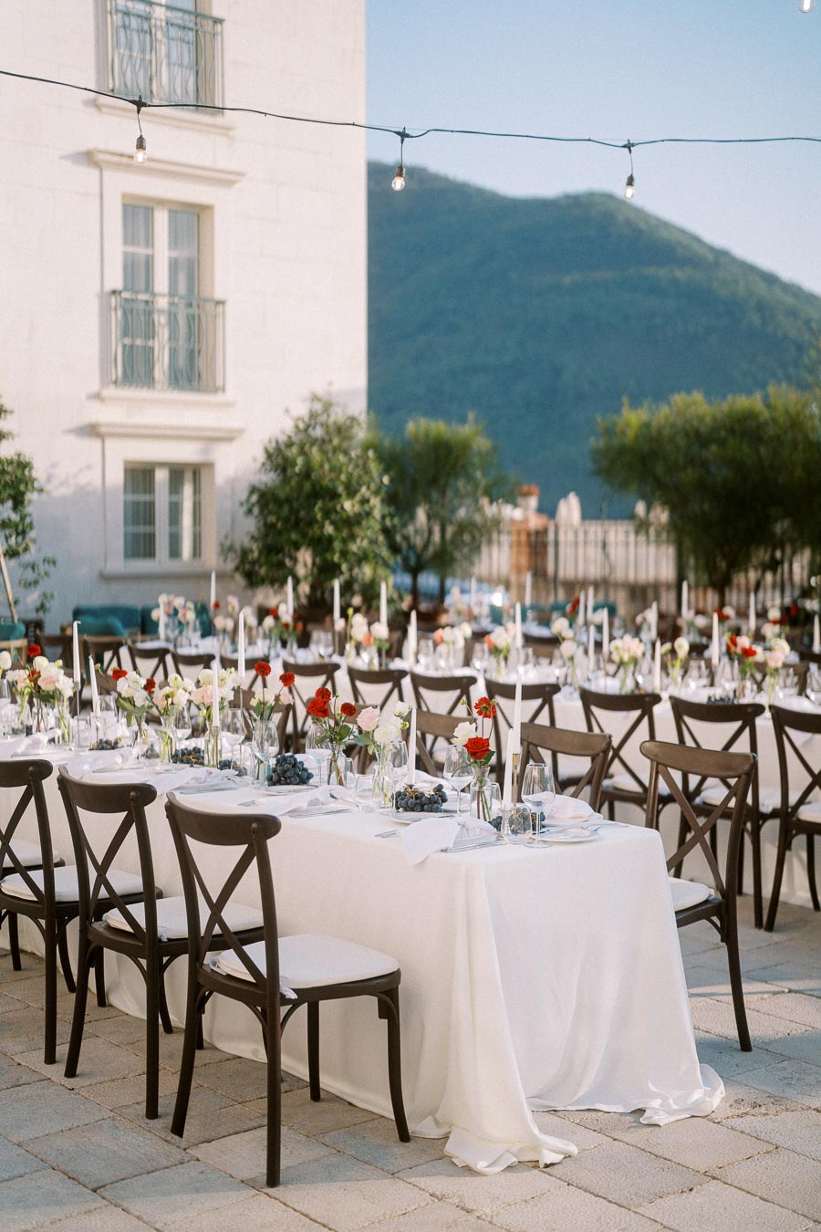 Outdoor wedding reception setup with elegantly arranged tables covered in white tablecloths, adorned with colorful flowers and candles, set against a scenic mountain backdrop.