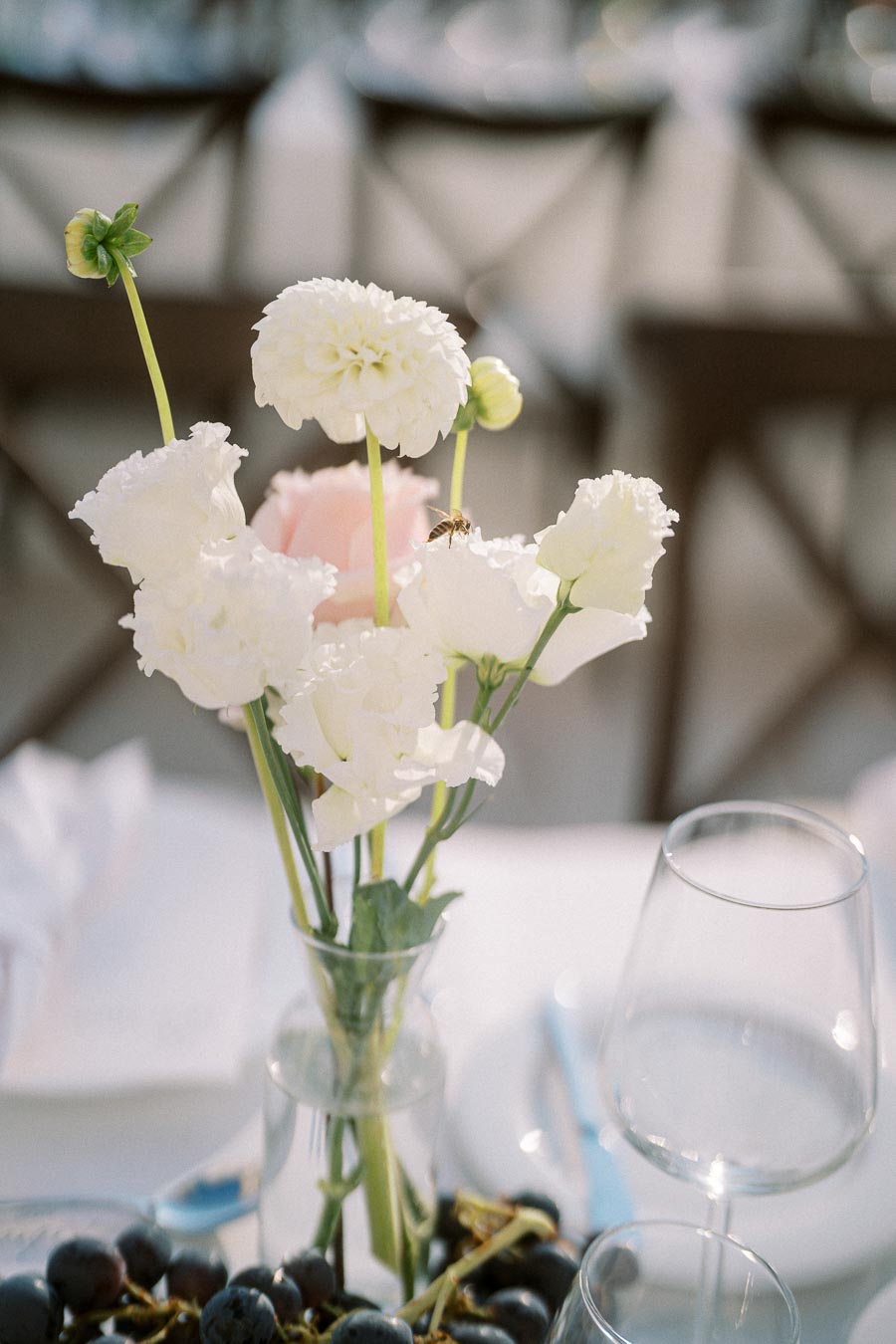 Elegant table setting featuring a delicate arrangement of white flowers in a glass vase, accented by a bee, set against a backdrop of wine glasses and grapes, creating a sophisticated atmosphere.