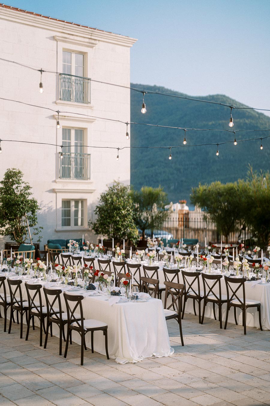 Outdoor wedding reception setup with elegantly decorated tables and chairs, surrounded by string lights and greenery, against a picturesque mountain backdrop.