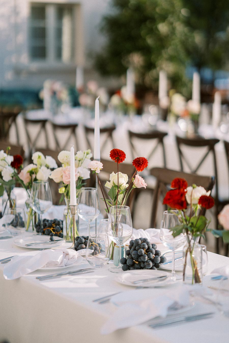 Elegant outdoor dining setup featuring a long table with white linens, adorned with red and white flowers in clear vases, tall white taper candles, wine glasses, and bunches of fresh grapes, creating a sophisticated and inviting ambiance.