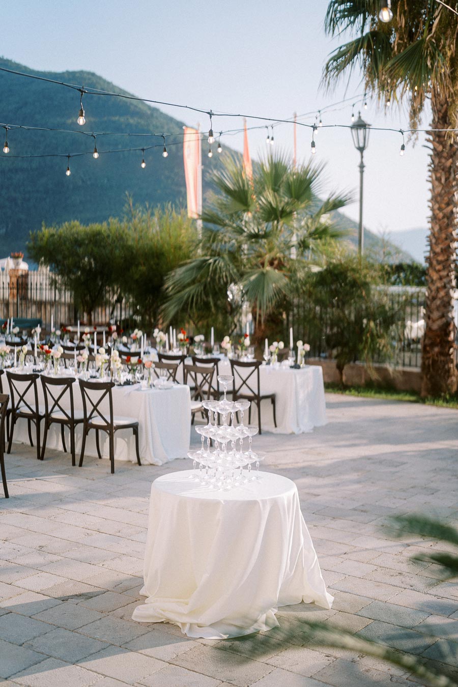 Elegant outdoor wedding reception setup with champagne glass pyramid on round table, surrounded by candle-decorated dining tables, palm trees, and string lights, with mountain view in the background.