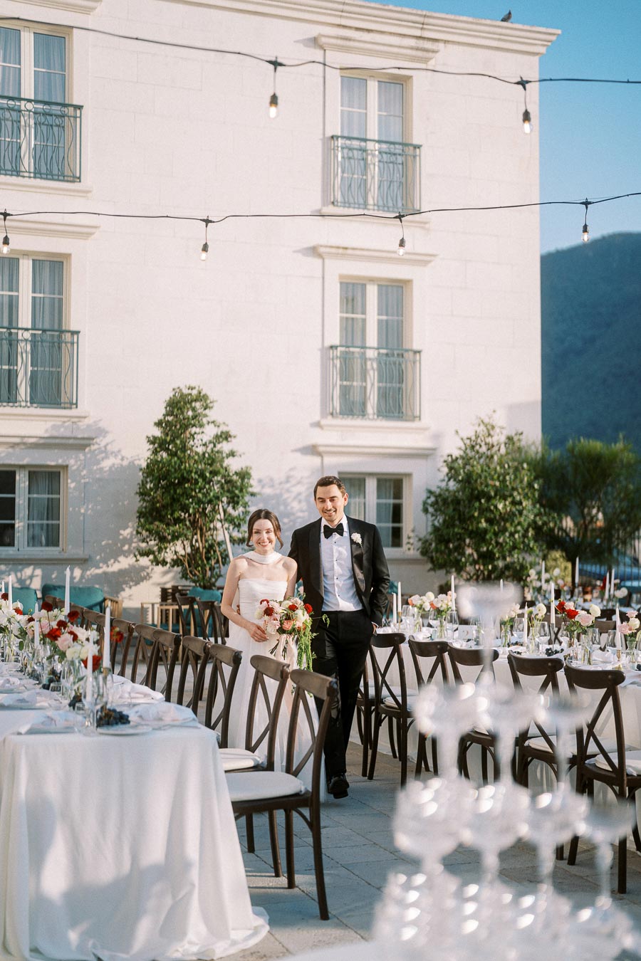 Elegant outdoor wedding reception with happy couple walking between beautifully decorated tables, floral arrangements, and softly lit ambiance.
