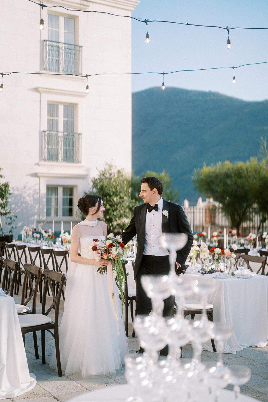 Elegant outdoor wedding ceremony with bride in white gown holding bouquet and groom in tuxedo, set against picturesque mountain and venue background, decorated tables and string lights.