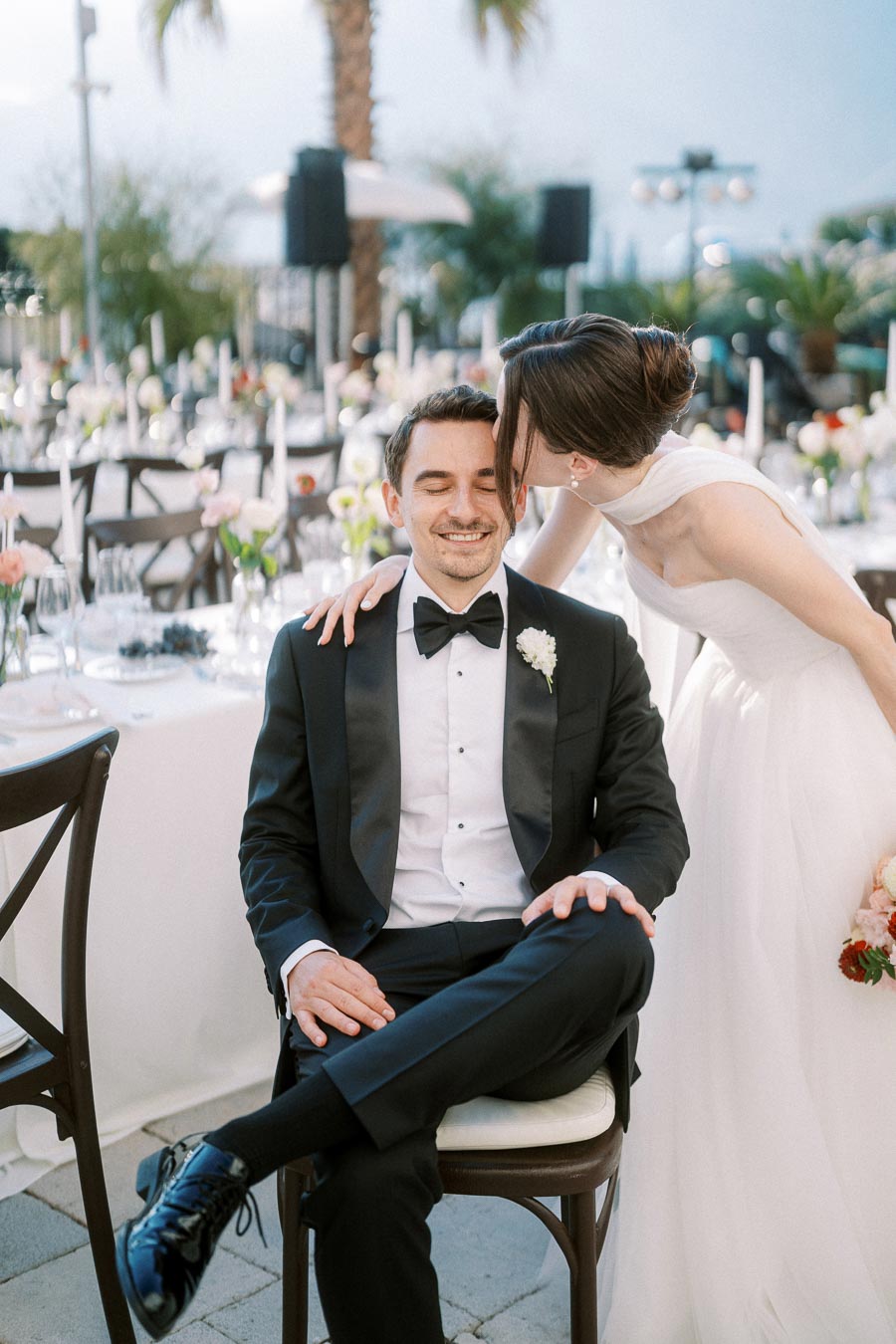 A bride in a white gown lovingly kisses a groom in a black tuxedo on the forehead at an elegant outdoor wedding reception with floral decorations and palm trees.