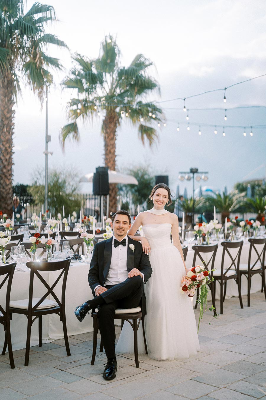 Elegant outdoor wedding setting with bride in white gown and groom in black tuxedo, posing by decorated tables under palm trees and string lights.