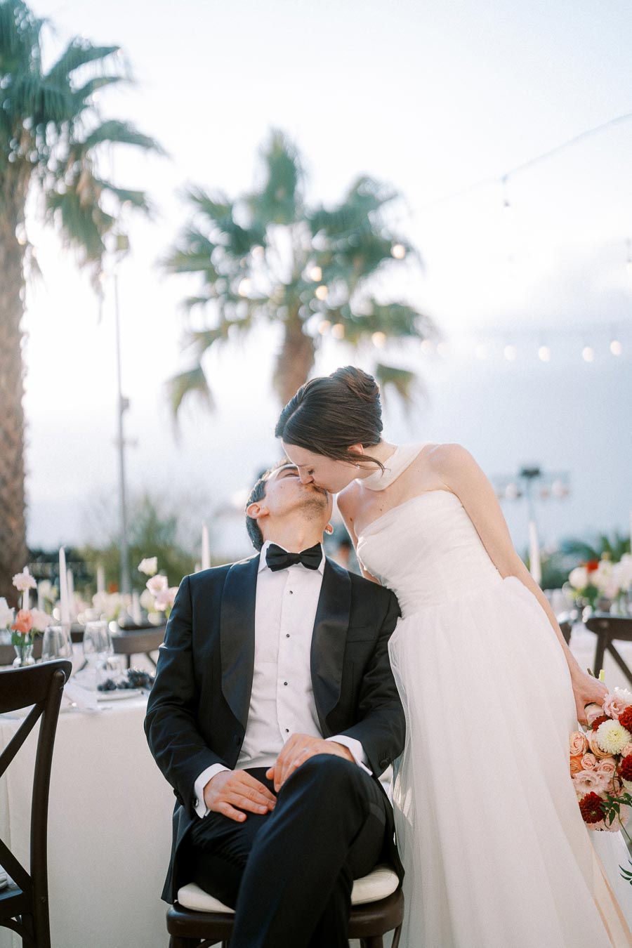 A bride in a white gown and groom in a black tuxedo share a romantic kiss at an outdoor wedding reception, surrounded by elegant table settings and palm trees under a soft twilight sky.