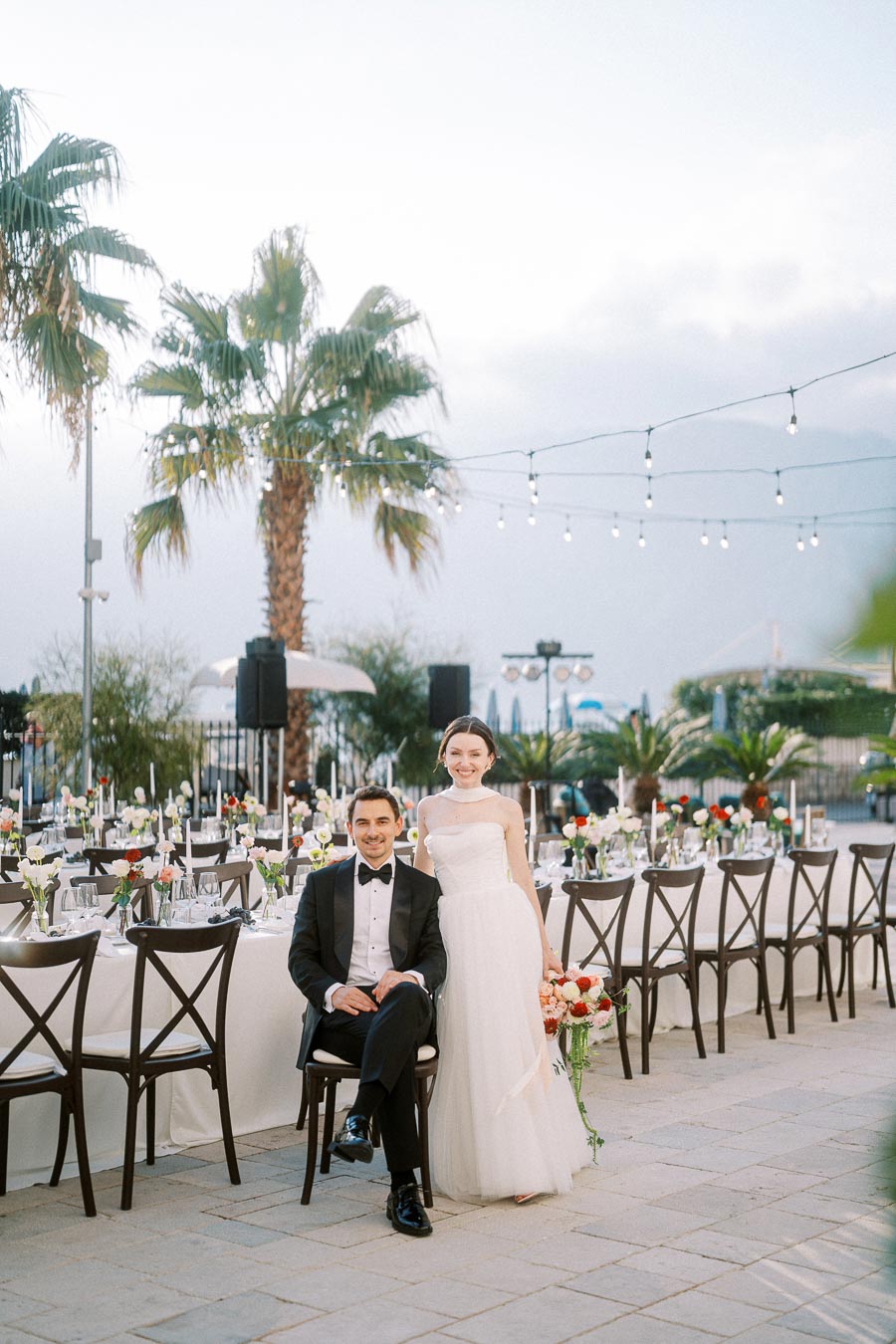 Outdoor wedding reception with a bride in a white gown and groom in a black tuxedo, posing happily next to an elegantly set banquet table adorned with flowers and under string lights, framed by palm trees.