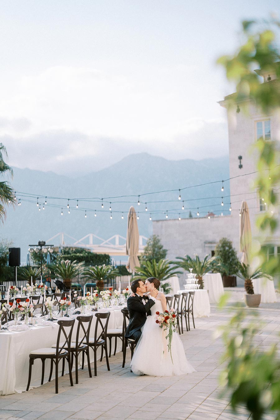 Wedding couple kissing at beautifully adorned outdoor reception, featuring elegant tables with flowers, surrounded by picturesque mountains and decorative string lights.