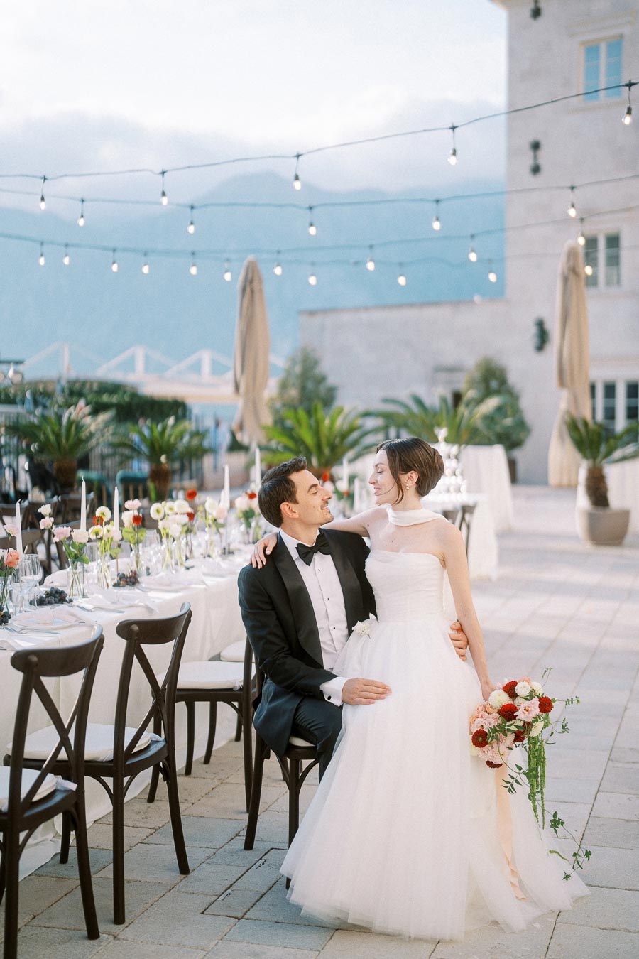 A newlywed couple sits lovingly together at an elegantly decorated outdoor wedding reception, adorned with string lights and floral centerpieces, in a picturesque setting with mountains in the background.