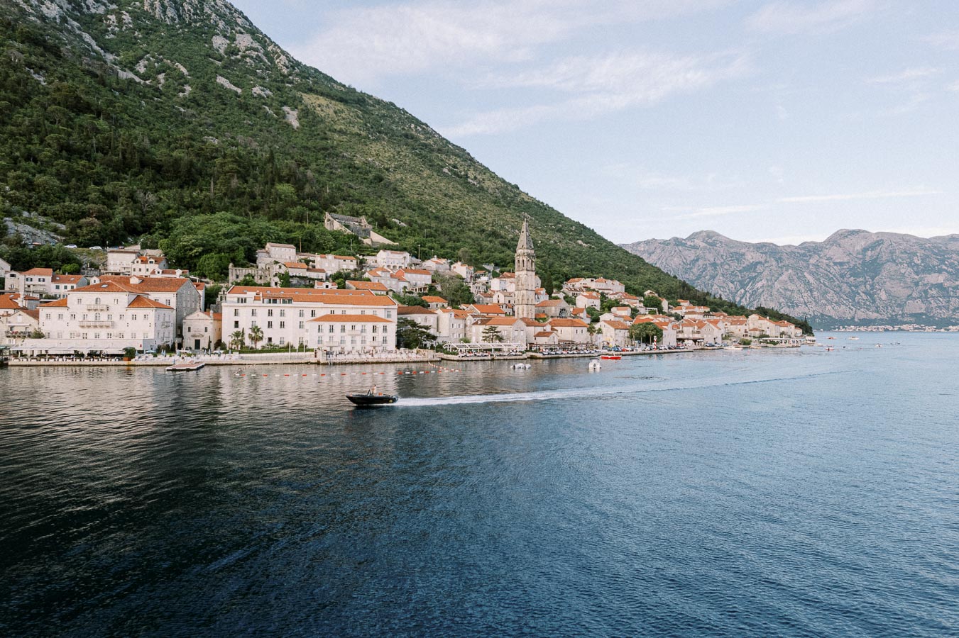 Scenic view of a coastal village with traditional Mediterranean architecture, featuring red-tiled roofs and a historic church tower, set against a backdrop of lush green hills and a serene blue sea. A motorboat speeds across the water, enhancing the picturesque landscape.