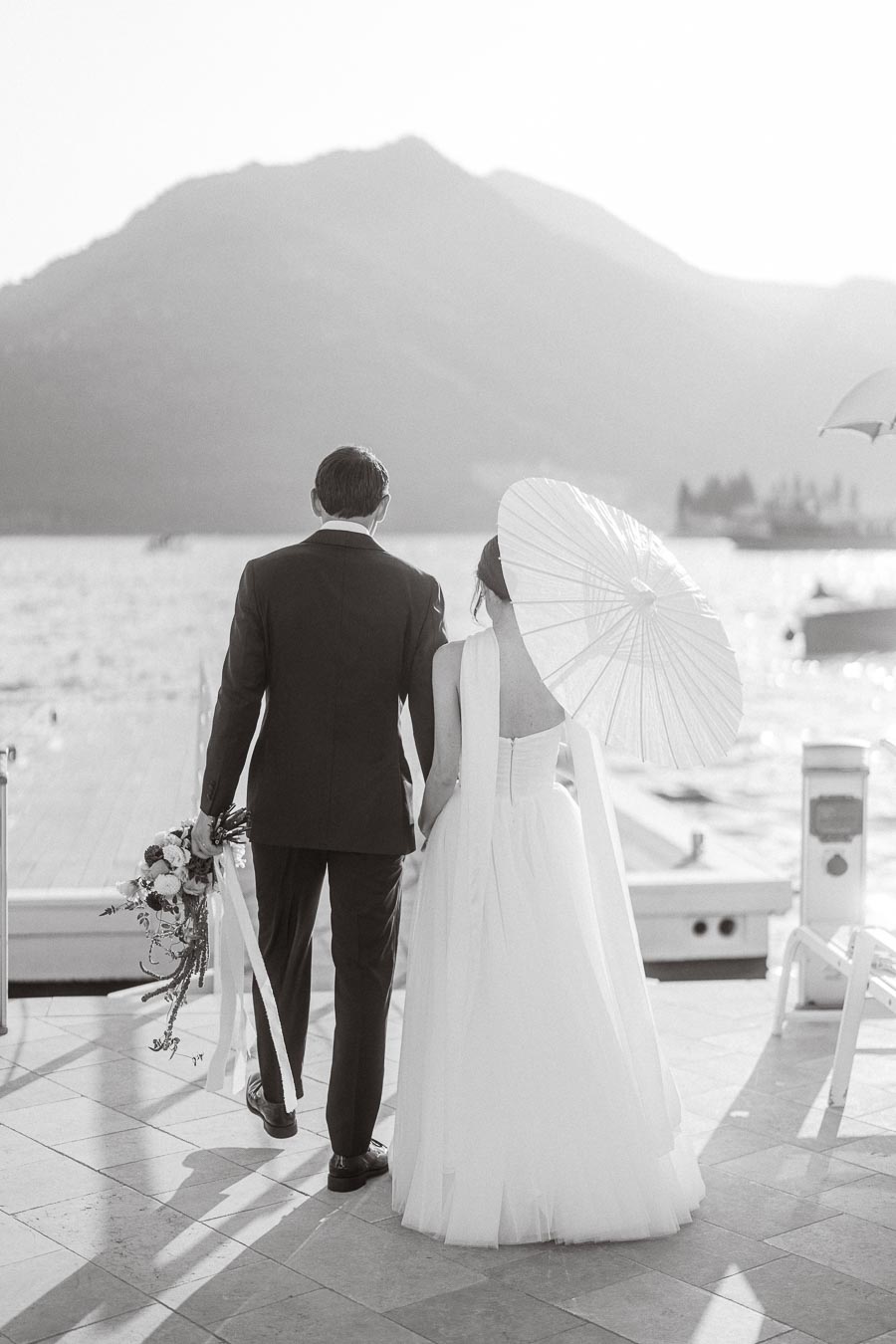 A couple in elegant wedding attire, with the groom holding a bouquet and the bride holding a parasol, walks towards a scenic lake with mountains in the background, captured in a black and white photograph.