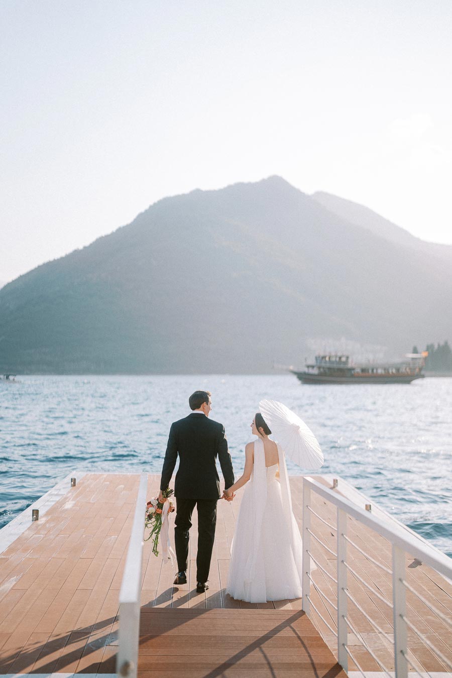A bride and groom holding hands on a wooden dock overlooking a serene lake, with mountains in the background and a boat in the distance, creating a picturesque wedding scene.
