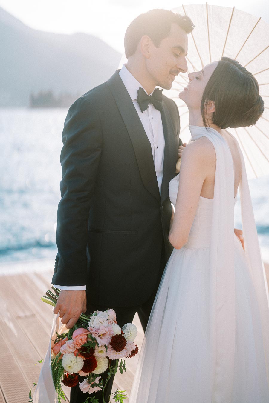 Elegant couple embracing on their wedding day, the groom in a black tuxedo and the bride in a white gown holding a colorful bridal bouquet, with a scenic waterfront backdrop.